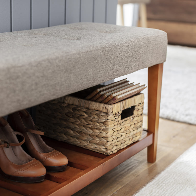 Banqueta gris con patas de madera con espacio de almacenaje. En ella hay un zapato color camello y un canasto de fibra natural con cuadernos.