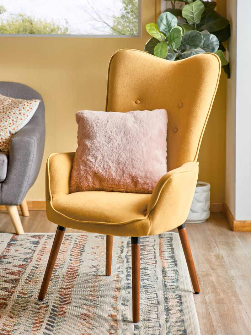 Poltrona con respaldo alto y con tapizado capitoné color mostaza. Tiene patas de madera. También tiene un cojín peludo rosa claro.