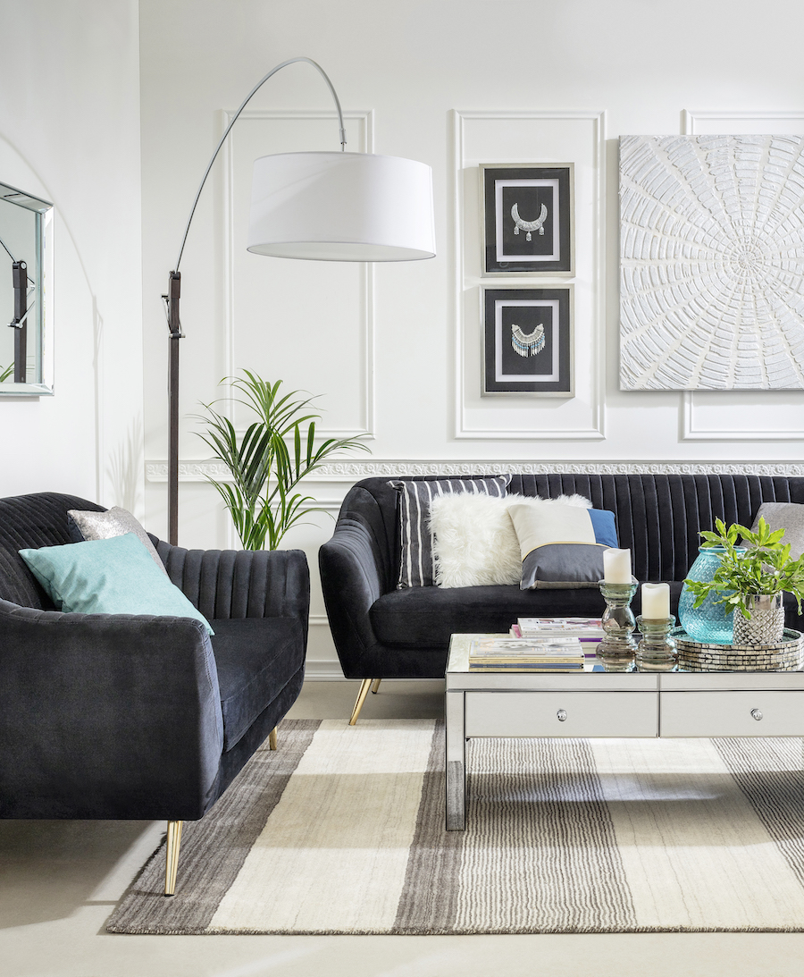 Sala de estar con 2 sofás negros de terciopelo, sobre ellos hay cojines blancos, grises y celestes. Al medio hay una mesa de centro espejada con velas, un florero y una planta encima. En la pared del fondo hay un gran cuadro blanco con textura.