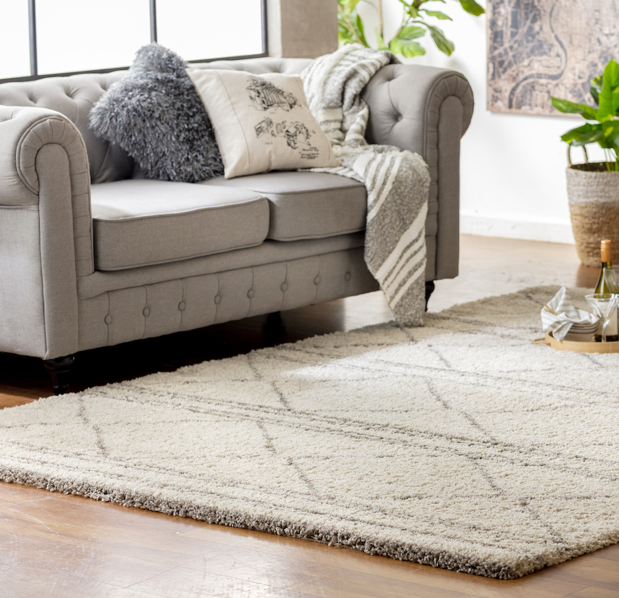 Sala de estar con un sofá capitoné gris claro. Sobre él hay un cojín peludo gris, un cojín crema con diseño y una manta gris con líneas blancas. Al frente hay una gran alfombra peluda color crema con líneas café claro.