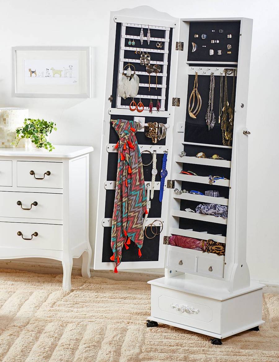 Dormitorio de estilo clásico con una cómoda y un mueble espejo blanco, cuya puerta está abierta y deja ver los distintos accesorios en su interior, como aros, anillos, collares, relojes y pañuelos.