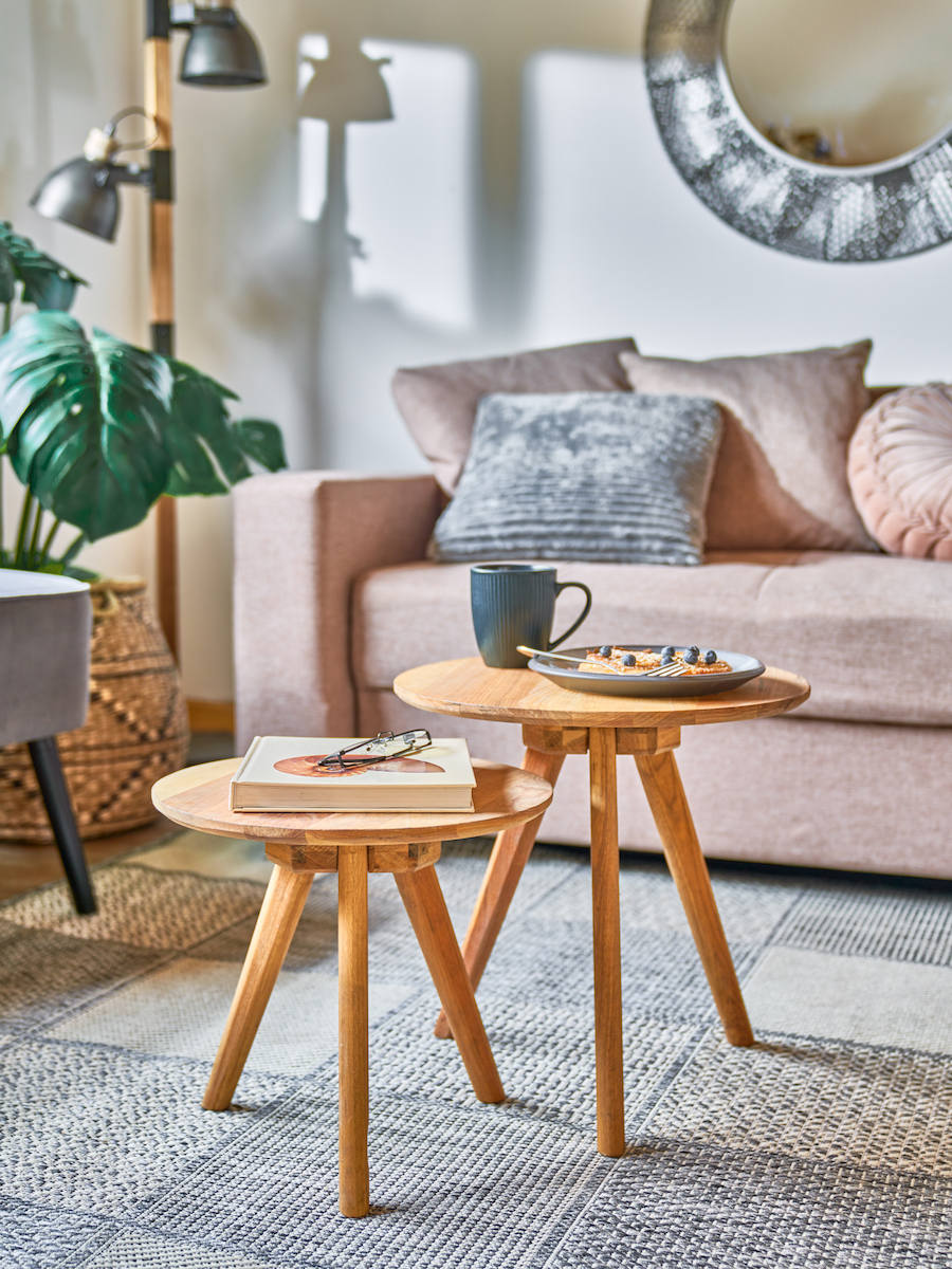 Sala de estar de estilo Boho chic con un sofá beige y cojines en tonos neutros sobre él, al costado hay una lámpara de pie y una planta. Adelante hay 2 mesas auxiliares de madera con 3 patas cada una. Sobre ellas hay un libro, un tazón y un plato.
