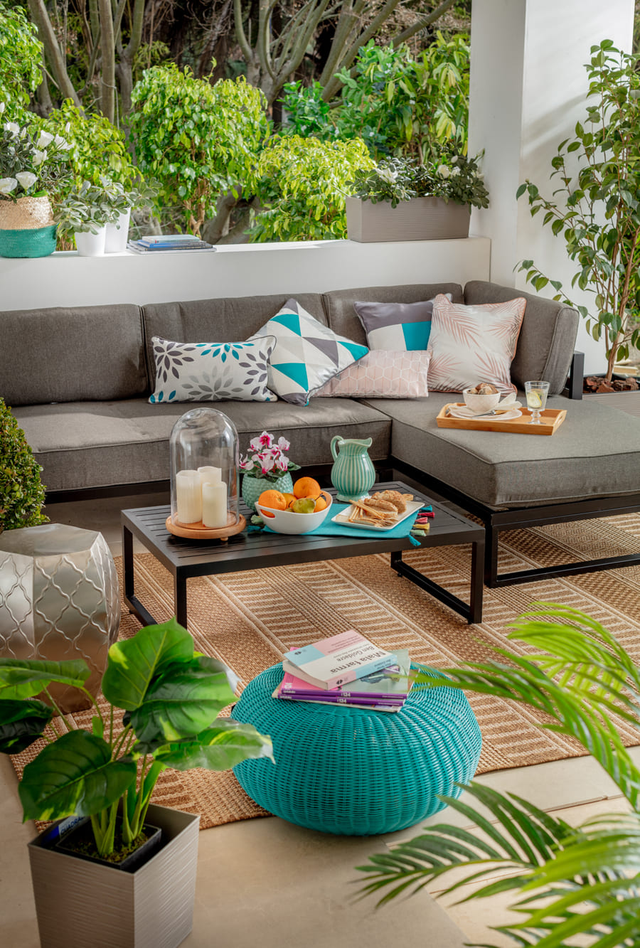 Una terraza con un pouf color calipso, un juego de living de terraza negro con cojines grises y una alfombra de fibra natural.