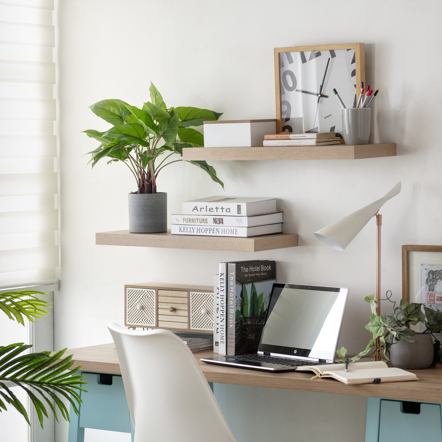 Home office con escritorio tipo caballete con base de madera color celeste. En cada pata hay un cajón. Base de madera sobre la cual hay un computador, artículos de escritorio y una lámpara moderna. Dos repisas flotantes de madera sobre muro blanco.