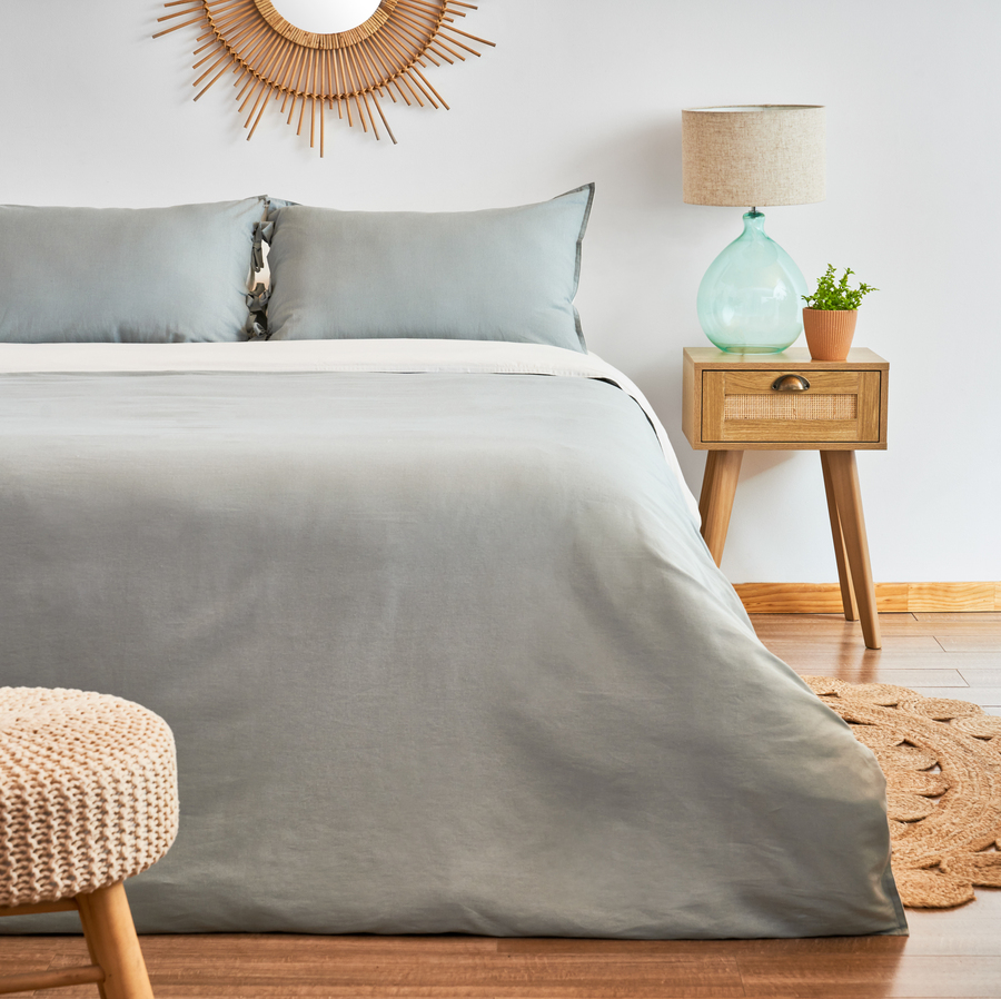 Dormitorio estilo boho, con cama bien hecha, con cubrecama y almohadas de color gris. Velador pequeño de madera, con un cajón. Sobre él hay una lámpara con base de vidrio y pantalla beige. Alfombra de yute de diseño floral.