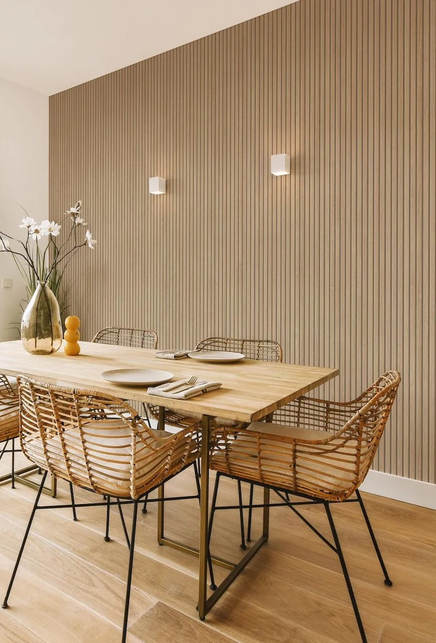 Comedor iluminado con luz natural, una de sus paredes es blanca y la otra tiene un panel decorativo de madera clara. La mesa de comedor también es de madera y las sillas, de mimbre. 