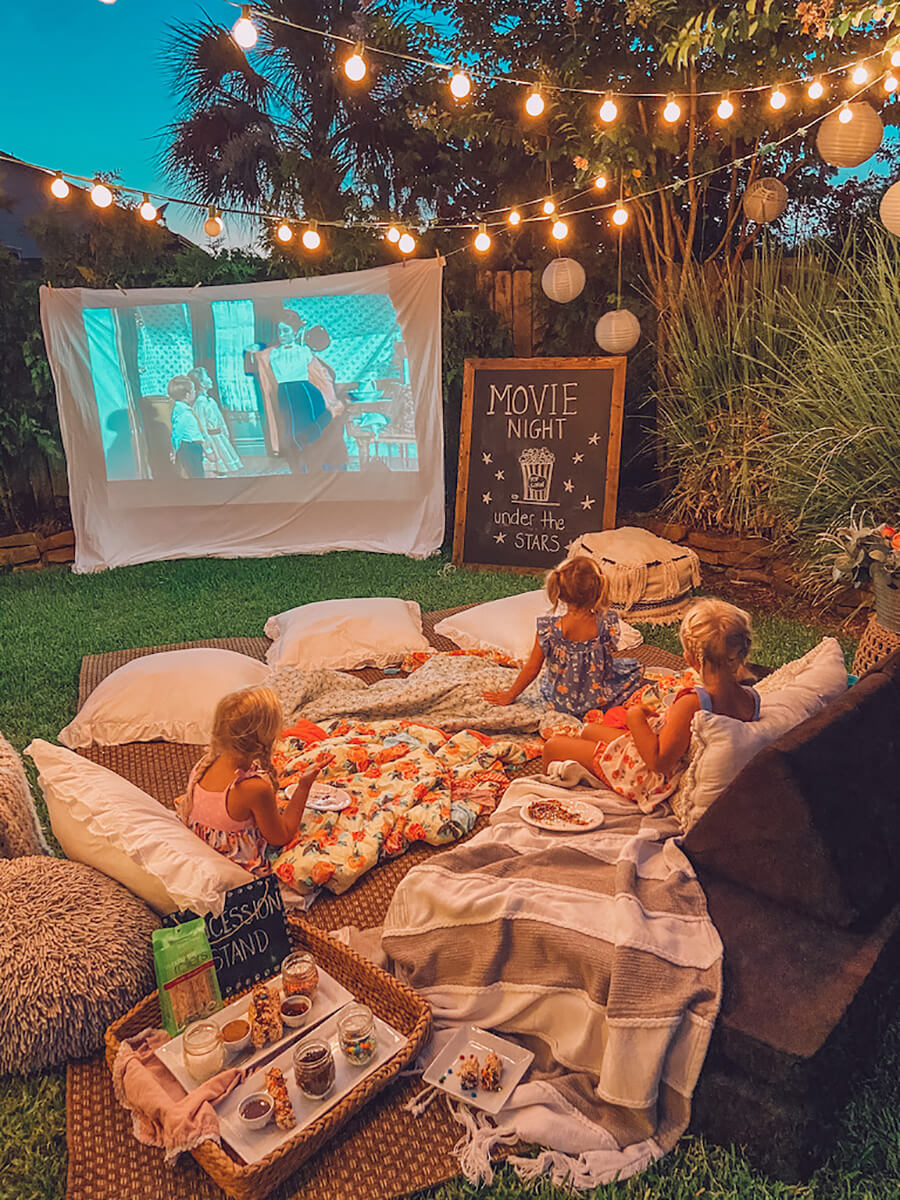 un jardín o patio ambientado para hacer una noche de cine en casa, con un proyector, unas mantas en el piso, cojines y unas niñas mirando una película.