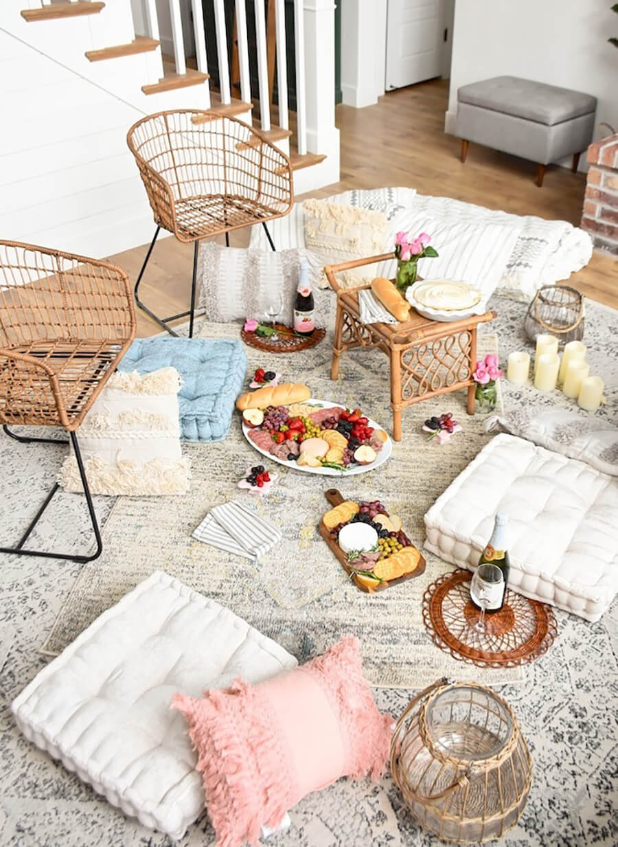una sala de estar o living ambientado para un picnic en casa, sobre una bella alfombra con cojines para sentarse, velas decorativas, ricos snacks y unas sillas y una mesa de centro de mimbre.