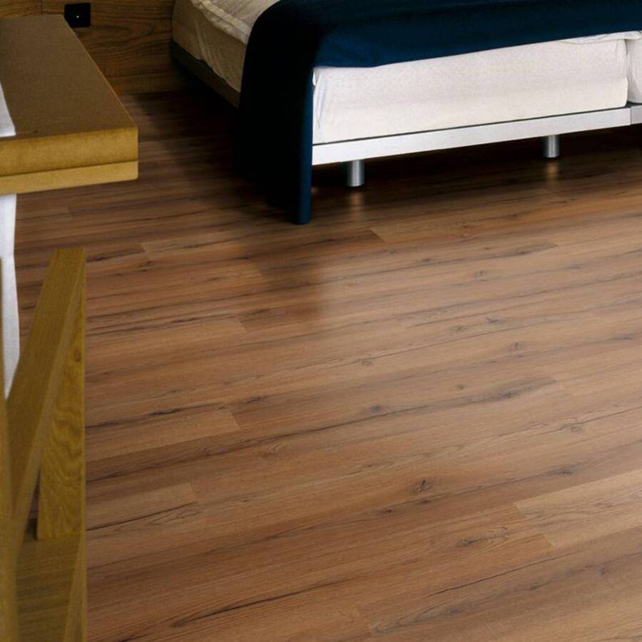 Detalle de piso flotante de madera oscura con vetas marcadas. Se asoma la esquina de una cama con colchón blanco y patas metálicas