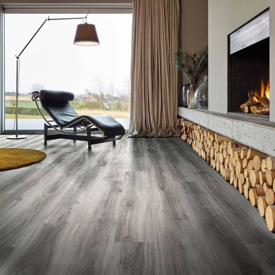 Living de estilo ejecutivo urbano. Piso vinílico de madera gris oscura o grafito. Chaise long de cuero negro y junto a él una lámpara de pie negra. Muro blanco con chimenea empotrada. Bajo ella hay un espacio para dejar leña.