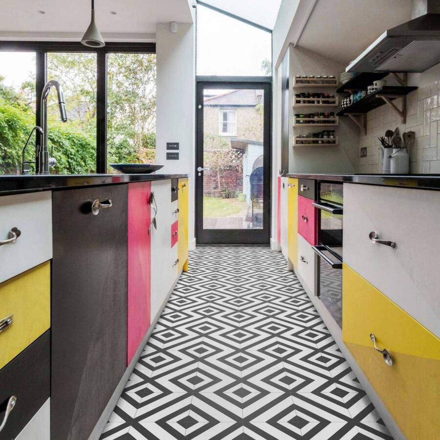 Cocina ecléctica con piso vinílico con diseño geométrico en blanco y negro. Muebles de diferentes colores, con cajones blancos, negros, amarillos y color sandía. Mesones color negro. Muros blancos y tragaluz alargado.