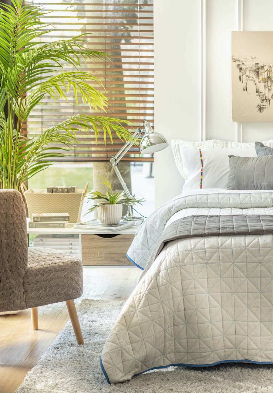 Dormitorio moderno con cama beige y gris. Ventana con persiana y gran planta tipo palmera. Sitial gris con patas de madera