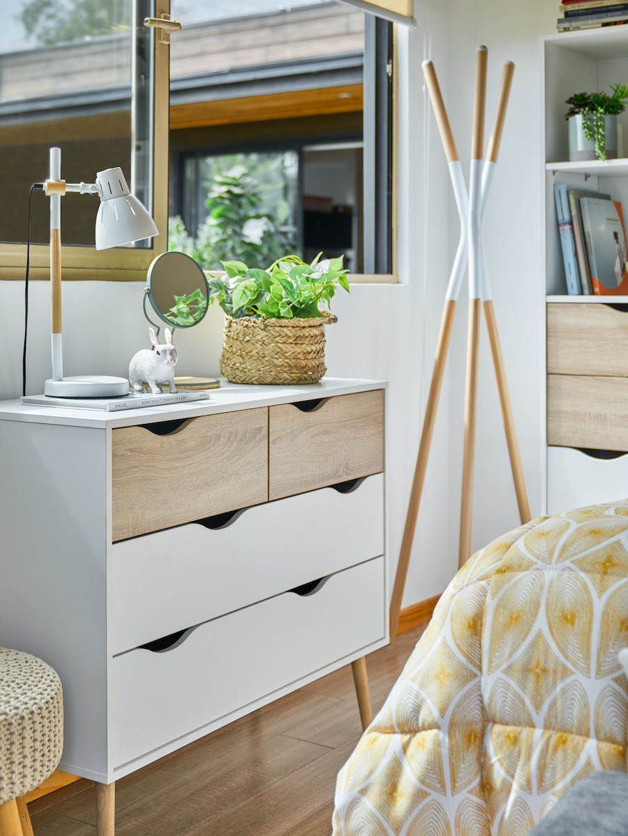 Dormitorio con cómoda blanca con cajones. Sobre ella hay un pequeño canasto de mimbre con una planta, un espejo y una lámpara de mesa nórdica.