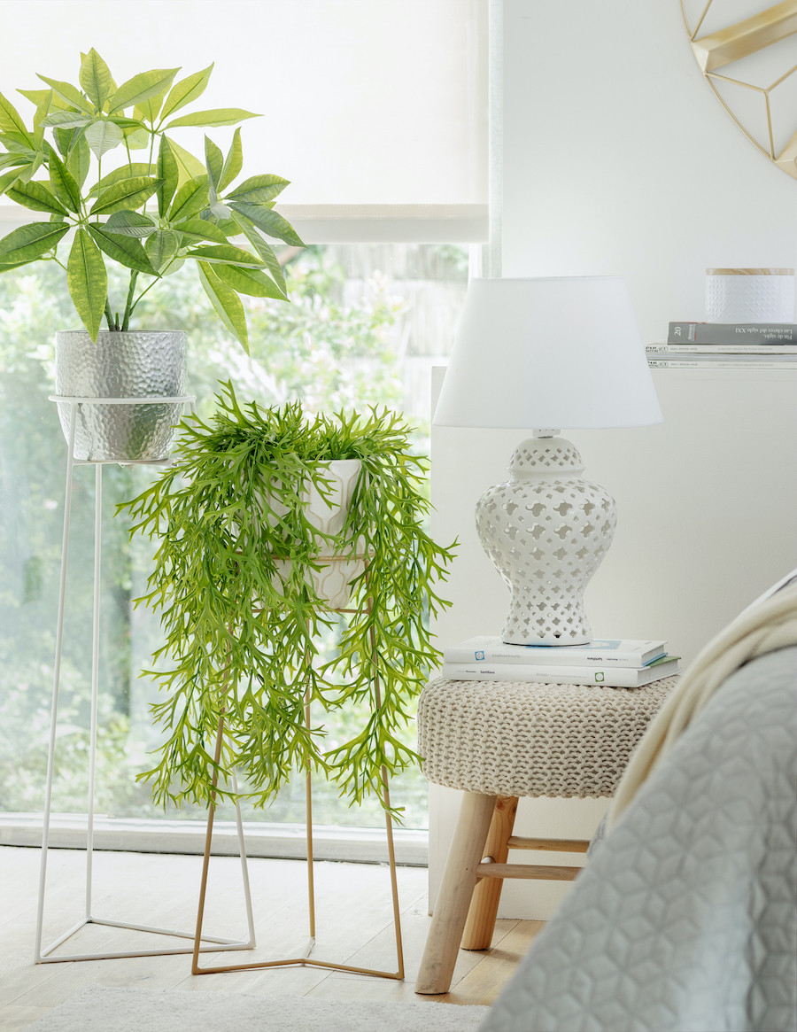 Costado de una cama con un pouf como velador. Al lado, dos plantas en unos pedestales metálicos y en maceteros blanco y gris.