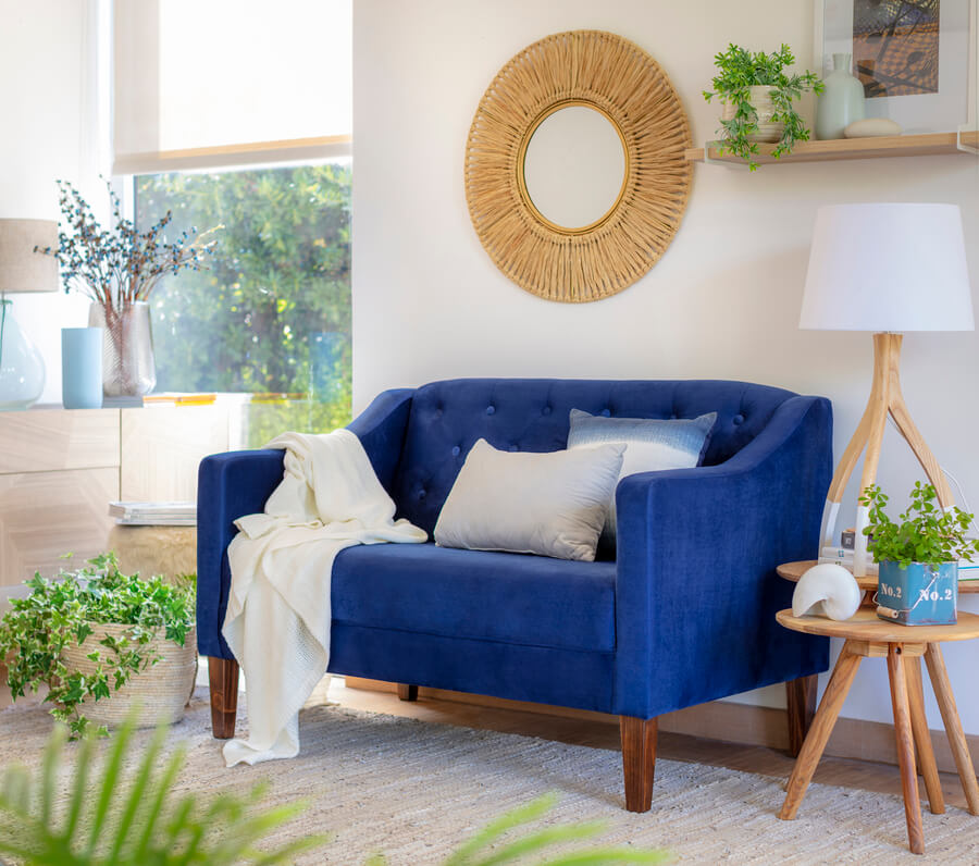 Living clásico con sillón de dos cuerpos azul. Dos cojines en tonos crema y azul y una manta blanca. Mesa de apoyo de madera, redonda, con lámpara de mesa. Espejo redondo con marco de mimbre. Repisa de madera flotante con cuadros y plantas