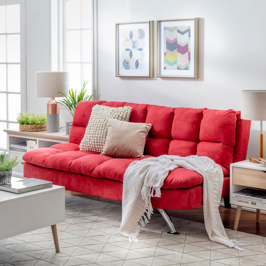 Living moderno con sofá cama color rojo y patas de metal. Sobre él hay dos cojines color beige y en el suelo hay una alfombra beige con líneas grises. Mesa de apoyo blanca con cajón de madera. Mesa de centro blanca con patas de madera