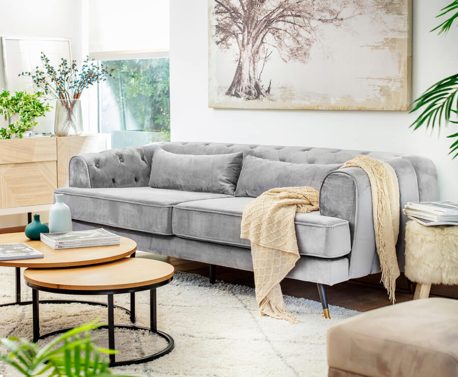 Living de estilo moderno, con sillón de líneas simples de color gris. Dos cojines grises y manta amarilla. Mesa de centro compuesta por dos mesas redondas de madera y patas metálicas negras. Alfombra beige con líneas grises. Gran cuadro en tonos beige