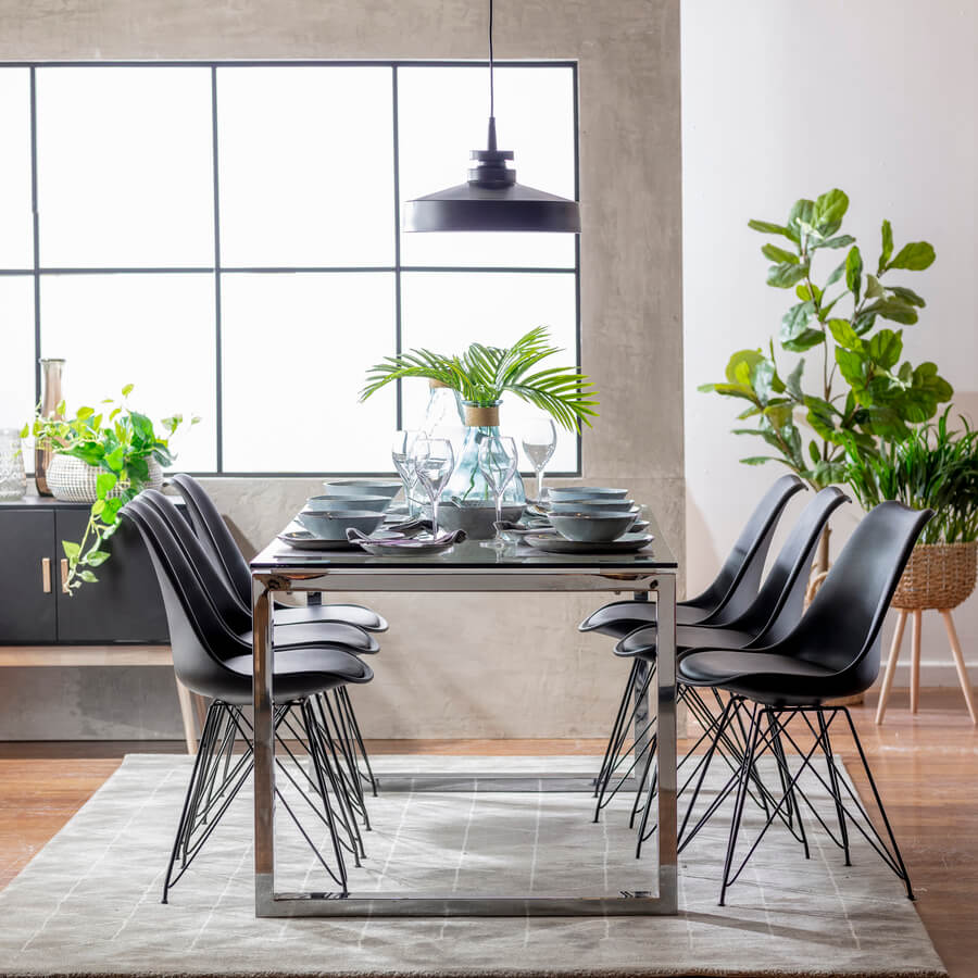 Comedor estilo industrial. Mesa rectangular de vidrio con patas metálicas. 6 sillas estilo eames, de color negro. Lámpara de techo negra, metálica. Gran ventanal con marco de fierro negro. Alfombra gris.