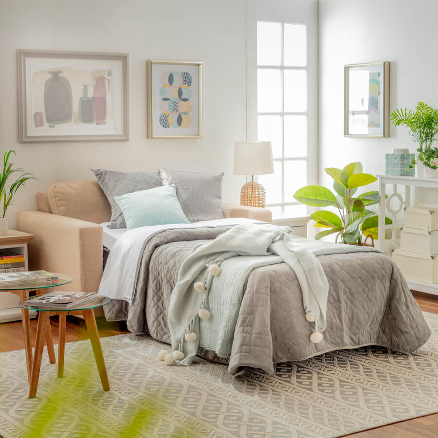 Living con sillón cama abierto. Cama armada con sábanas blancas, cubre cama gris y manta celeste. Alfombra grande con diseño geométrico. Muro blanco con dos cuadros sobre el sillón. Set de mesas de apoyo redondas.