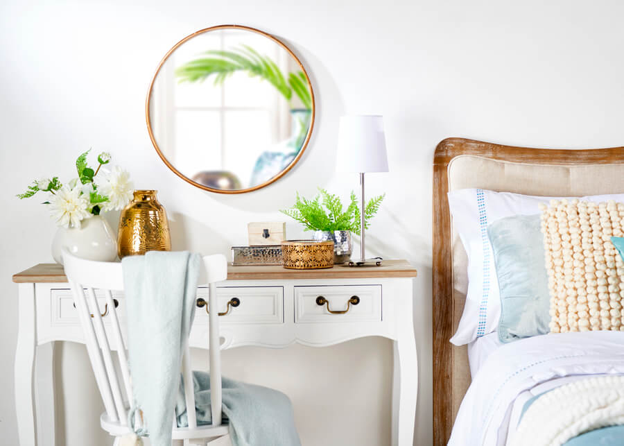 Dormitorio con escritorio de velador. Mesa blanca estilo provenzal, con tres cajones y tiradores metálicos. Silla blanca de madera con manta celeste. Espejo redondo con marco dorado. 
