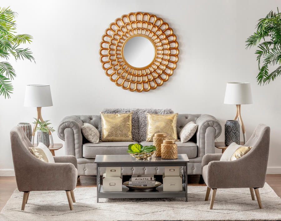 Living de estilo clásico. Sillón capitoné de tela, gris, con cojines dorados. Dos sofás de un cuerpo a cada lado, de color gris. Mesa de centro gris con dos niveles, con espacio de almacenaje donde hay cajas y libros. Dos mesas de apoyo con lámparas.