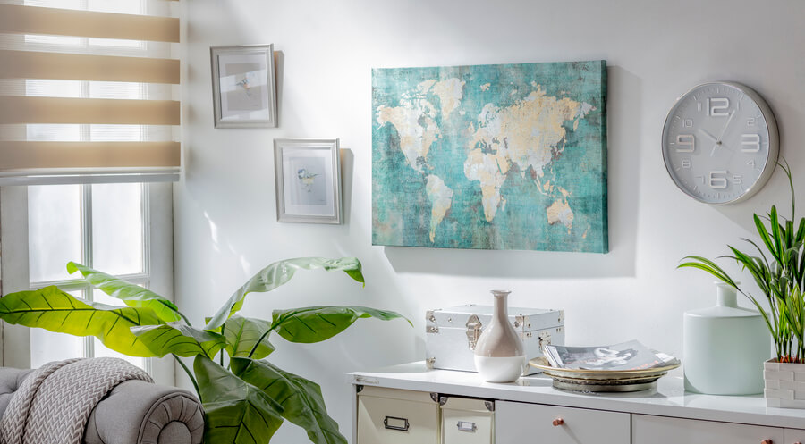 Esquina de una habitación. Cómoda blanca con repisas y cajones, sobre ella hay adornos y cajas organizadoras. En el muro hay un cuadro de un mapa, dos cuadros pequeños y un reloj. 