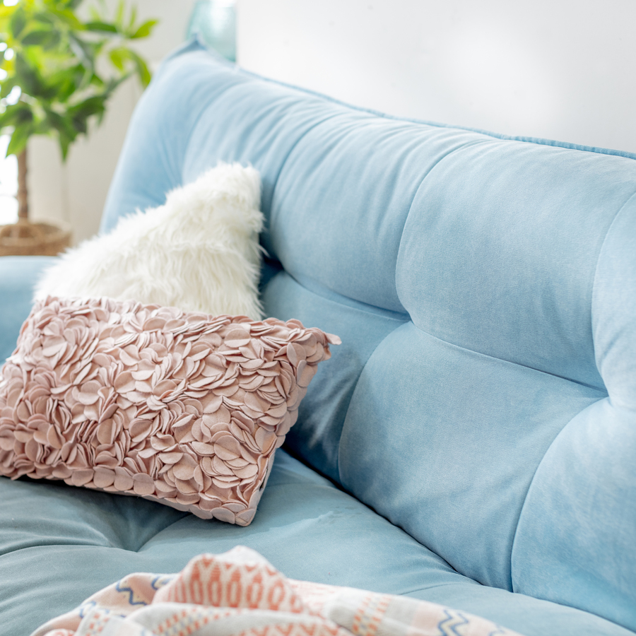 Detalle de sillón de color celeste, con tela suave aterciopelada. Un cojín con relieve tipo pétalos de color palo rosa y otro cojín de tela peluda, blanco. 