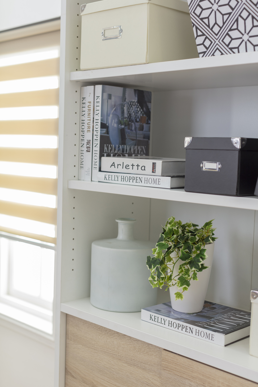 Repisa abierta, blanca, de melamina. Libros y una caja negra para almacenar. Jarrón blanco y una planta en un pequeño macetero blanco. Junto a la repisa hay una ventana con una cortina roller duo color crema.