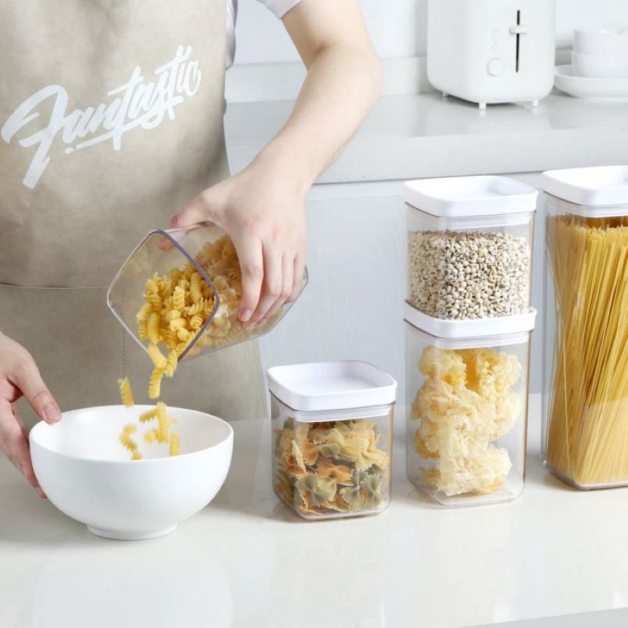 Set de 5 contenedores de acrílico con tapa blanca. 4 de ellos están apoyados en un mesón blanco. Uno de ellos los tiene una mujer en las manos y lo está vaciando en un bowl blanco.