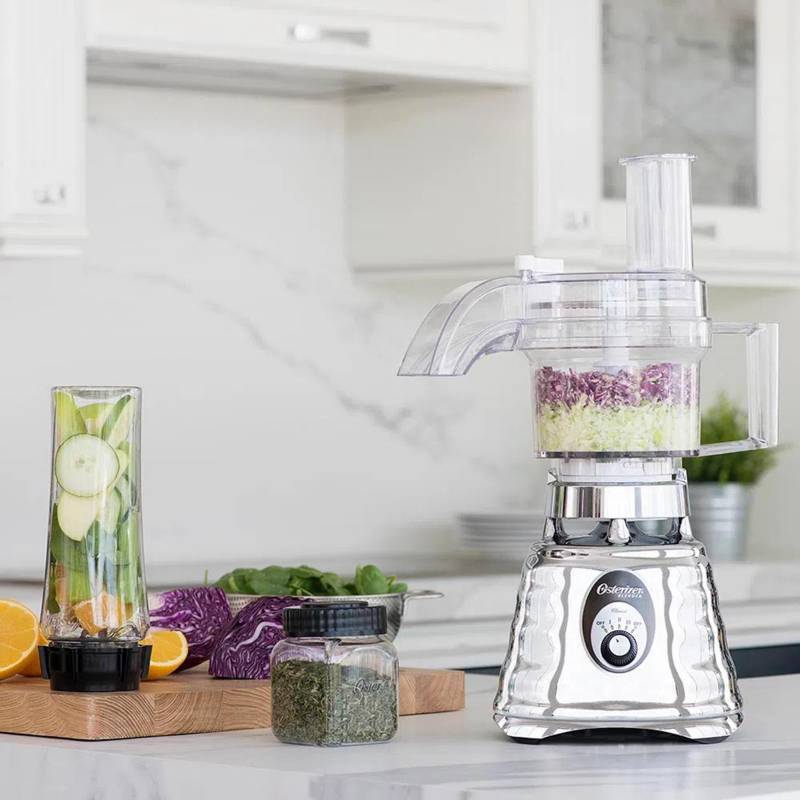 Procesadora de alimentos con con hojas picadas de repollo, pepino y otras verduras. La máquina está sobre un mesón de mármol blanco y al lado hay una tabla de madera con más verduras.