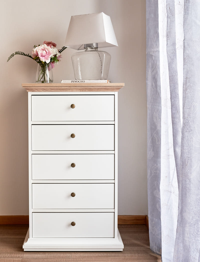 Cajonera estrecha blanca con cinco cajones. Su cubierta es de madera y sostiene una lámpara de mesa blanca con base transparente y un jarrón blanco con flores.