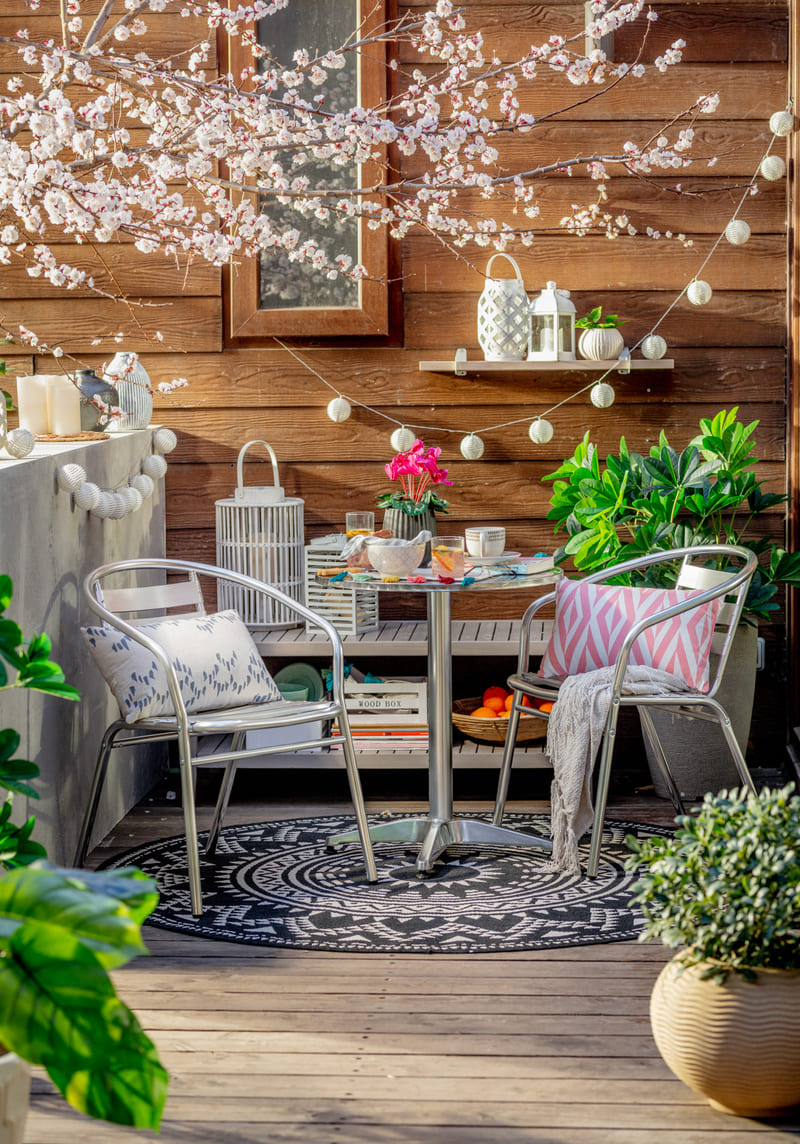 Balcón con un juego de balcón para dos de metal, que están sobre una alfombra redonda con patrones blanco y negro. Al fondo hay una banqueta que sostiene libros y portavelas. En la pared hay una repisa y un marco con una ilustración.