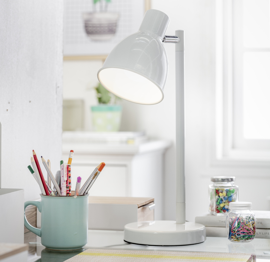 Lámpara de mesa blanca, metálica con la luz encendida. A su lado hay una taza celeste con lápices, frascos con clips y pins de colores. Atrás se ve un mueble blanco y un cuadro de plantas