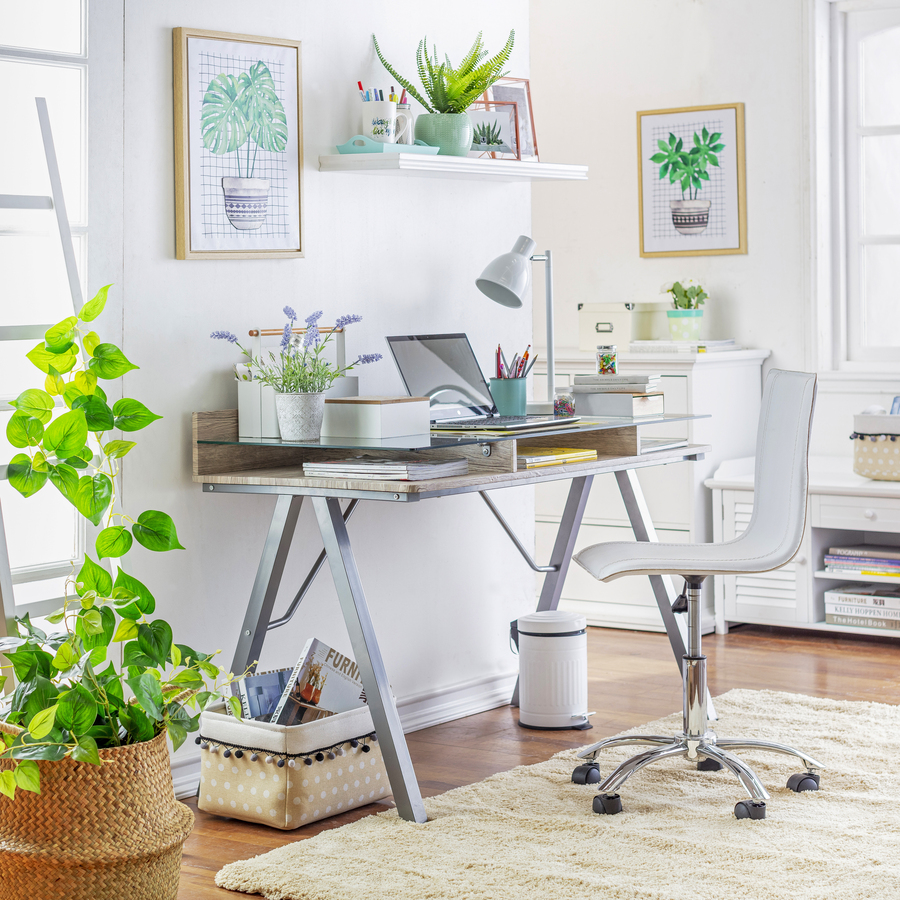 Escritorio moderno y luminoso. Mesa de metal, madera y vidrio, silla con ruedas blancas, Sobre el escritorio hay artículos de oficina, organizadores y una lámpara. Repisa flotante blanca y cuadro de plantas. Canasto de mimbre en el suelo, alfombra beige