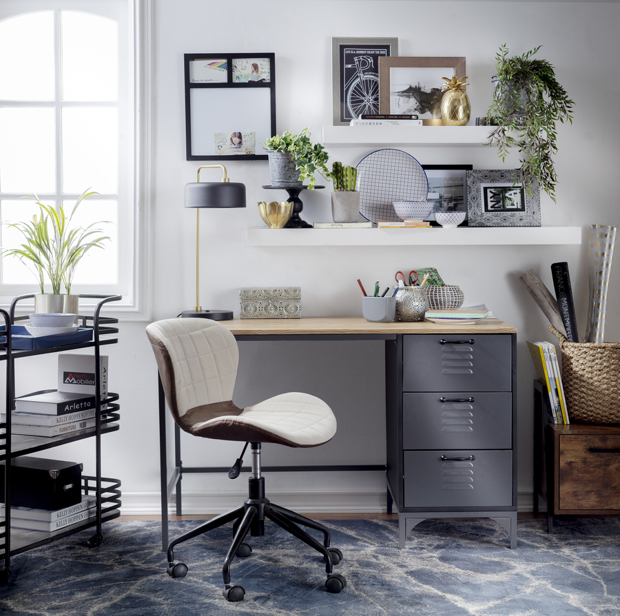 Escritorio de estilo industrial. Mesa de metal negra con tres cajones, base de madera. Silla con ruedas negra con beige. Alfombra gris. Dos repisas flotantes blancas con adornos, cuadros y plantas. Carro metálico negro de tres niveles.