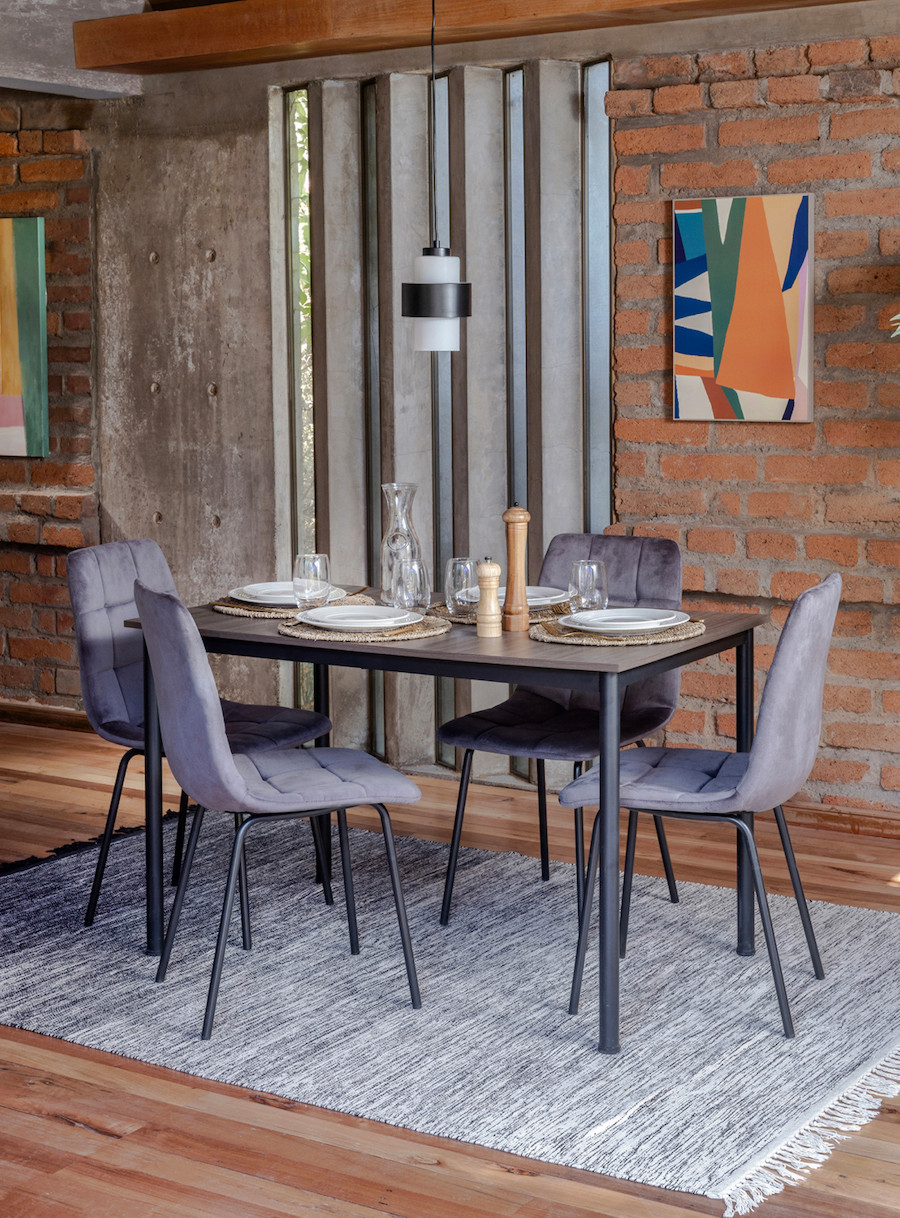 Comedor con una mesa de estructura negra, sillas grises, muros de ladrillo y alfombra gris.
