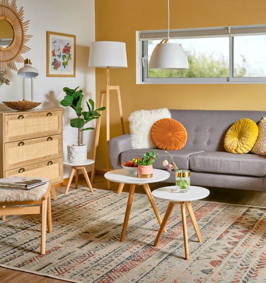 Living de estilo boho. Un muro color amarillo mostaza, y otro muro color blanco. Sofá de dos cuerpos, color gris, de estilo nórdico. Tiene 4 cojines, uno planco peludos, dos redondos de color amarillo y naranjo y otro estampado con flores. Set de dos mesas
