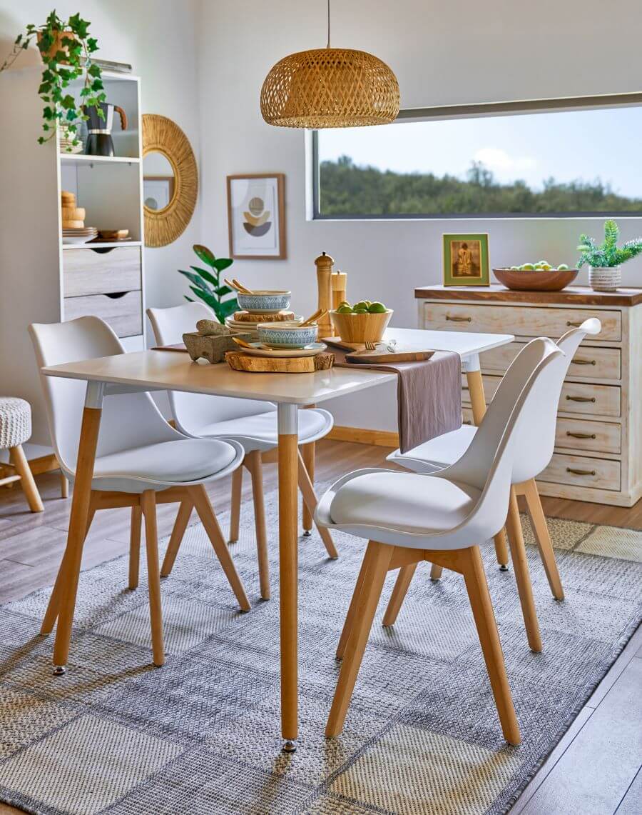 Comedor con mucha luz natural, muros blancos y ventana alargada en la parte superior. Comedor de base blanca con patas de madera, para 6 personas. Hay 4 sillas tipo "eames", con base blanca acolchada y patas de madera. Sobre la mesa hay vajilla de cerámica