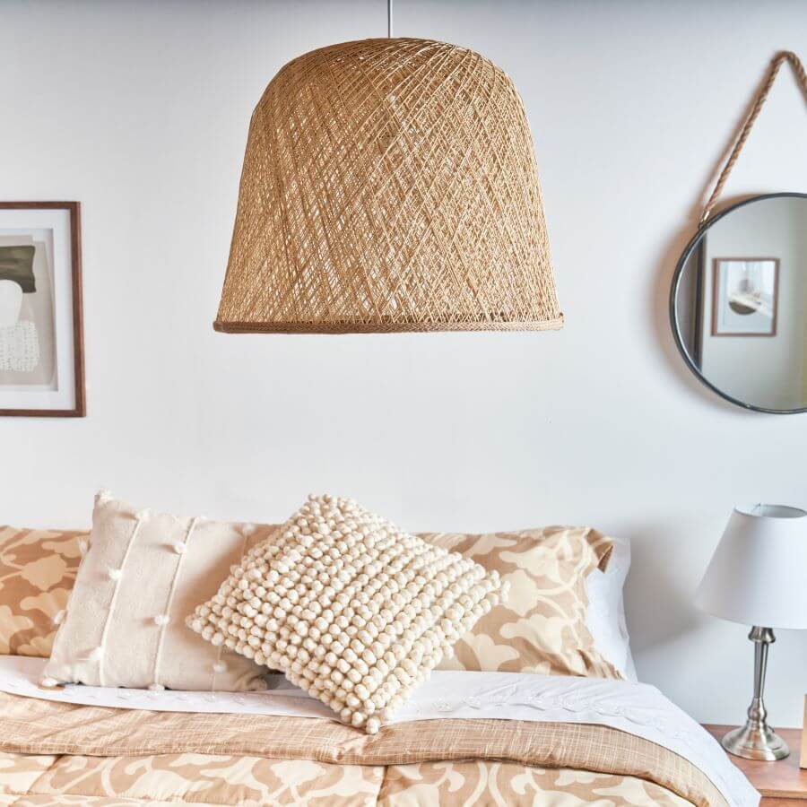 Dormitorio estilo boho. Gran lámpara de techo de fibras naturales en tonos café claro. Cama de dos plazas con cubrecama color beige y café claro, con dos cojines con textura. Muro blanco, con un cuadro y un espejo redondo con una cuerda para colgar. Junto