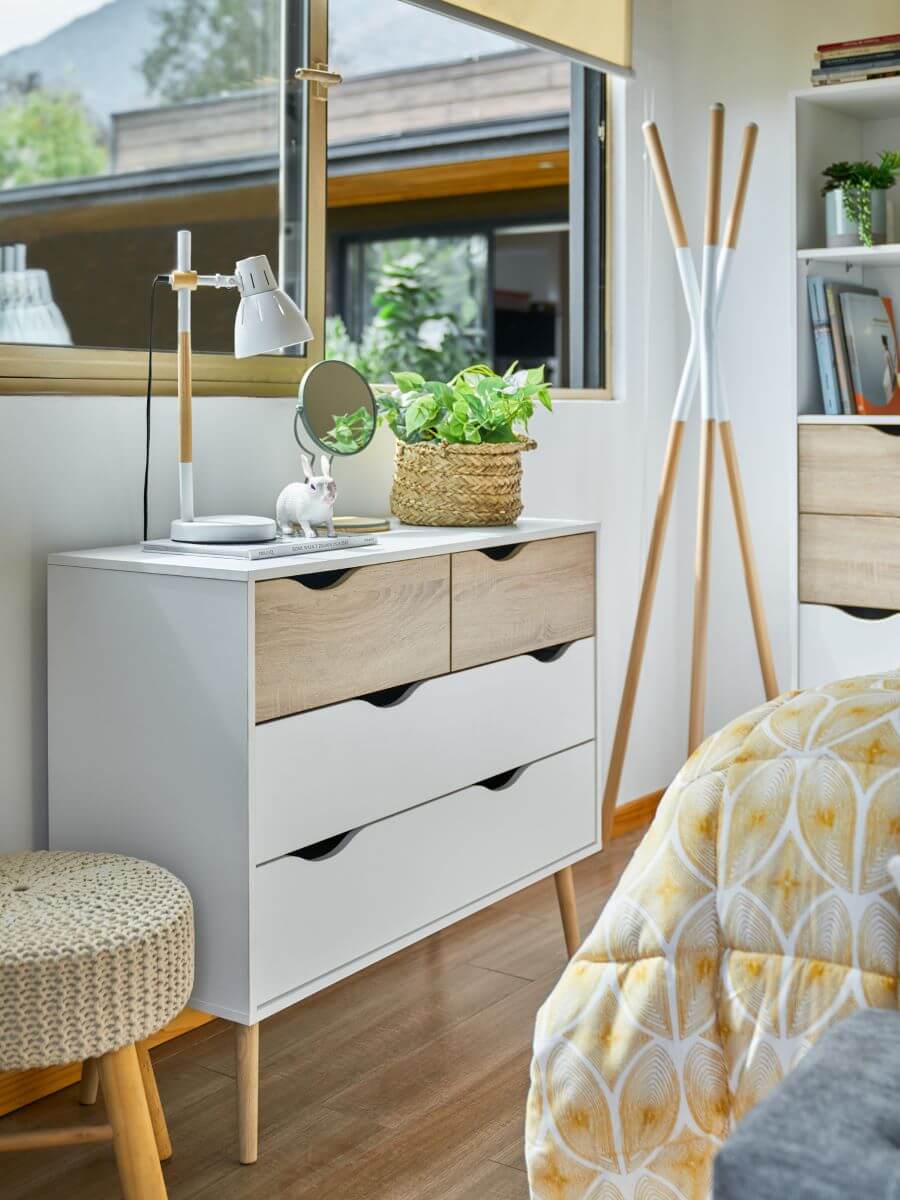 Dormitorio con muros blancos y gran ventana abierta. Mueble cajonera con dos cajones inferiores largos y dos cajones más pequeños en la parte superior. Es de melamina blanca y patas de madera. Sobre la cómoda hay una planta con un macetero de canasto de mi