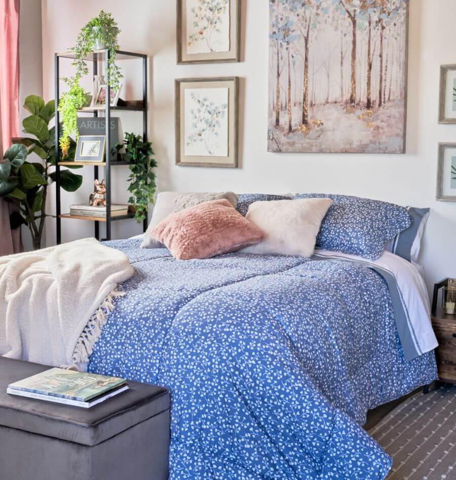 Dormitorio estilo clásico. Cama de dos plazas con plumín azul con pequeñas ramas blancas. Dos cojines blancos y uno rosado, peludo. A los pies hay una manta color blanco. Banqueta gris con revistas arriba. Sobre la cama hay una gran pintura de árboles en t