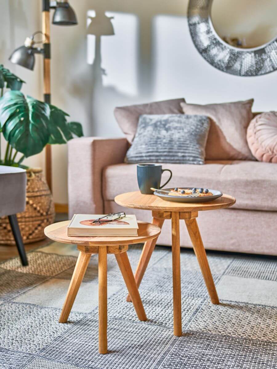 Detalle de dos mesas de apoyo de madera, redondas. Sobre ellas hay un plato, una taza y un libro. En el suelo hay una alfombra tipo "patchwork" con tejidos en tonos grises. Atrás se de un sofá de color rosa oscuro, con cojines del mismo tono y uno gris.